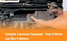 A mechanic holding a catalytic converter in front of a car engine, showing what a catalytic converter looks like and how it connects to a vehicle’s exhaust system.