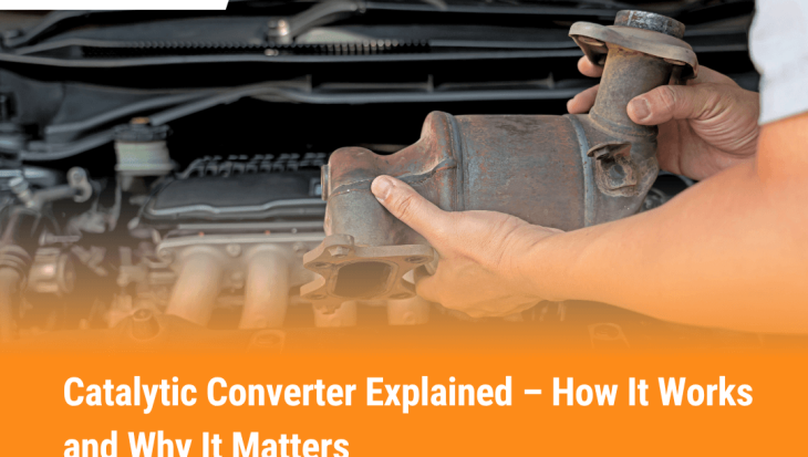 A mechanic holding a catalytic converter in front of a car engine, showing what a catalytic converter looks like and how it connects to a vehicle’s exhaust system.